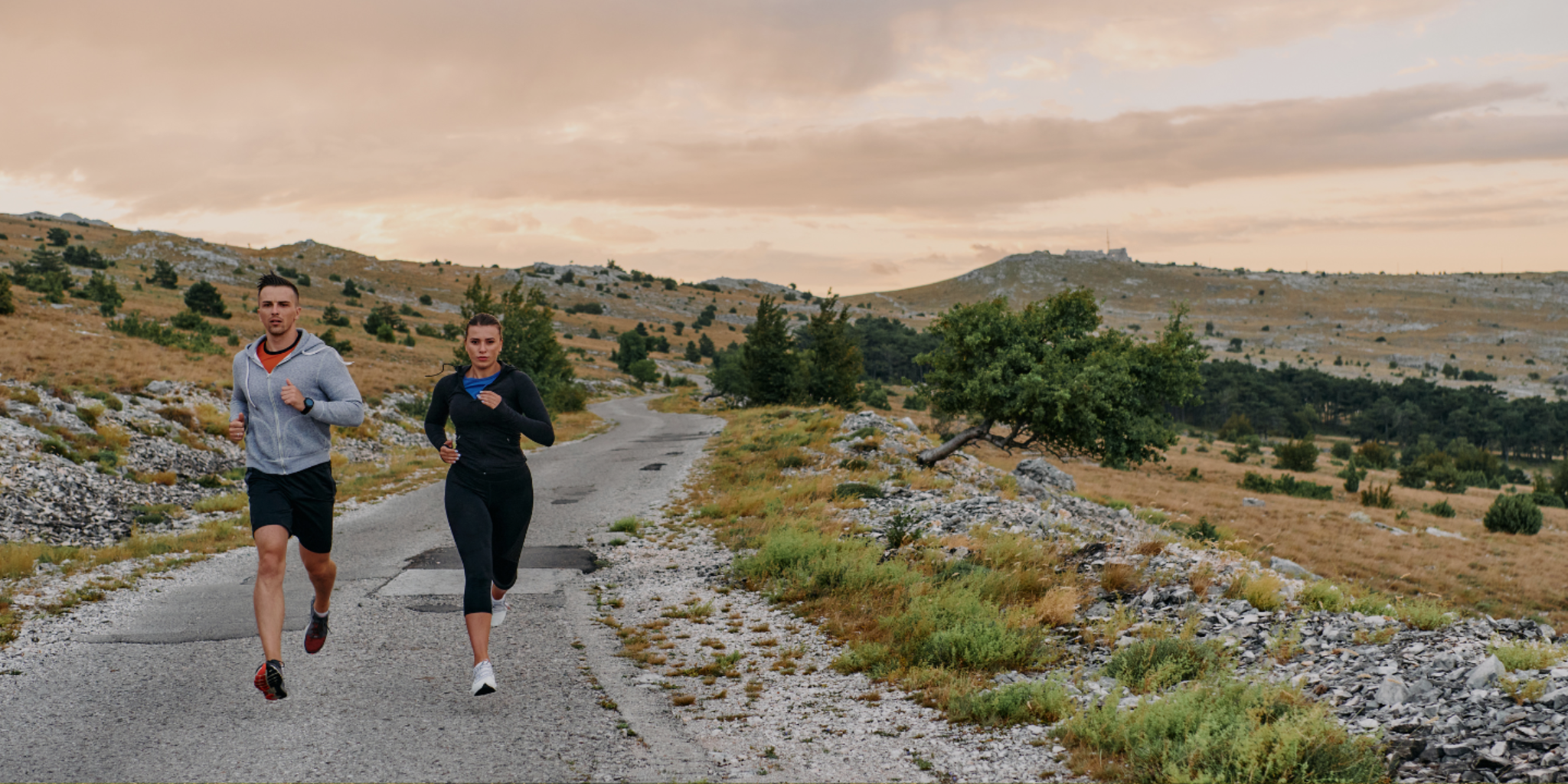 Two people running on a path in a scenic landscape with mountains and trees.
